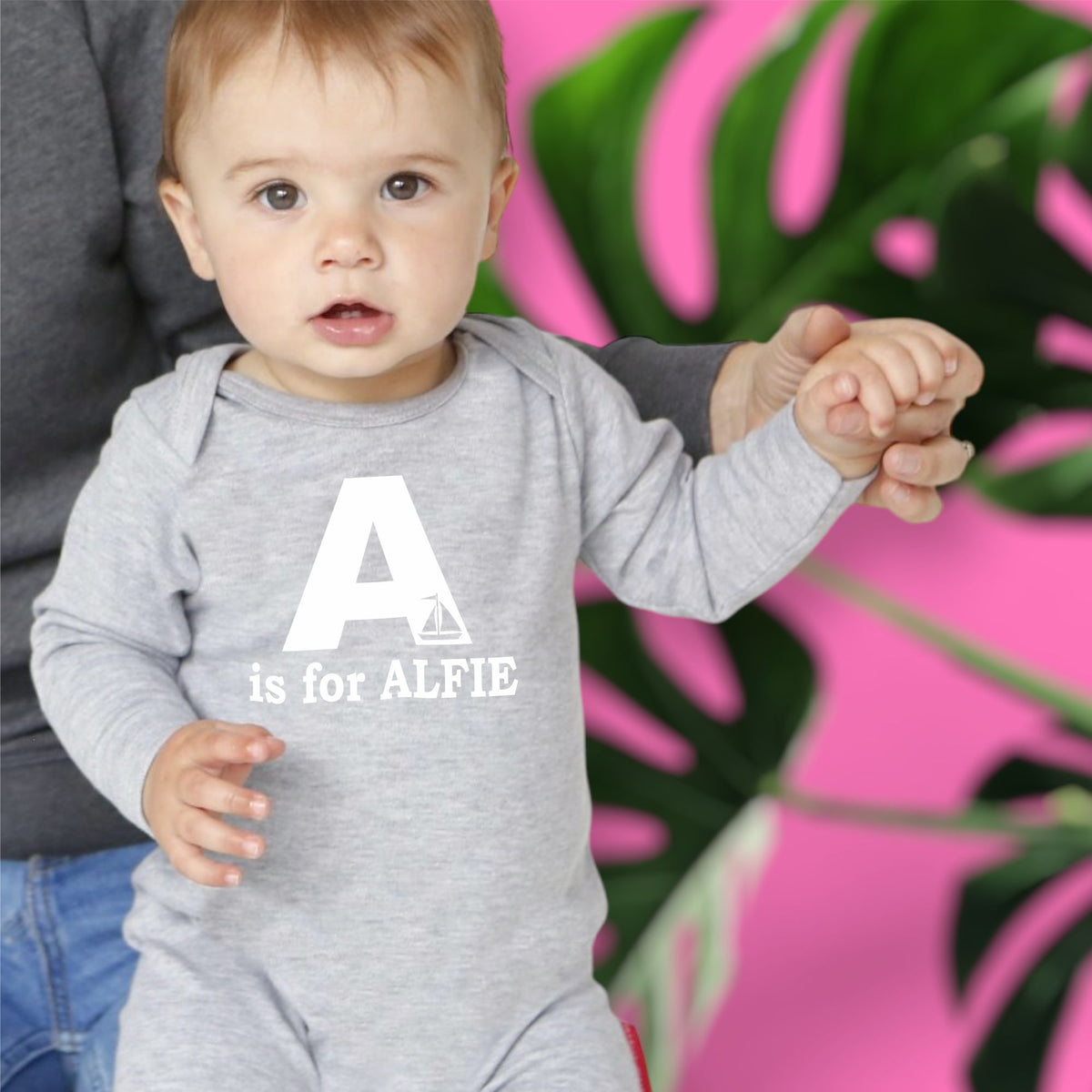 Baby wearing a grey onesie with 'A is for ALFIE' text, standing against a pink and green leafy background.