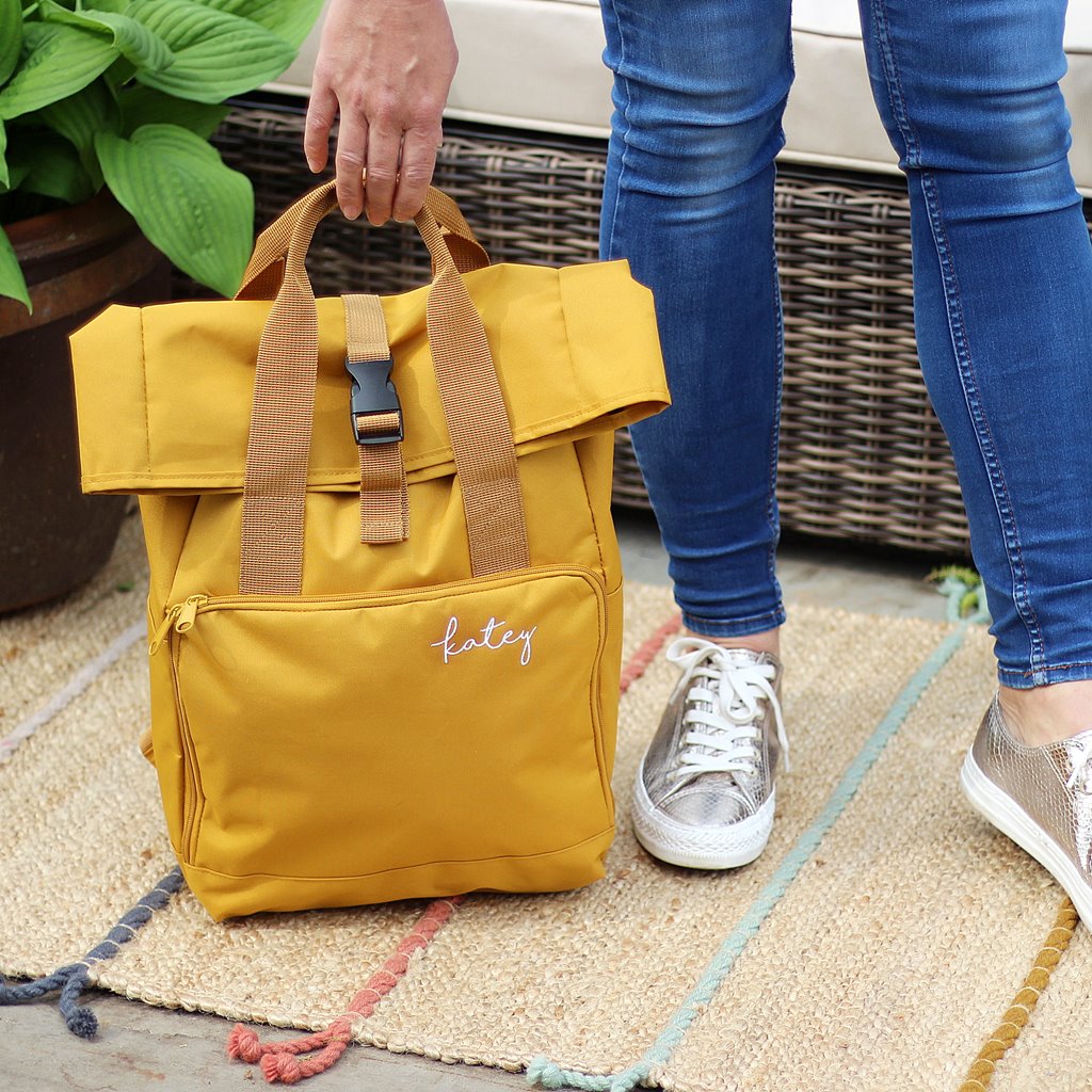 Yellow backpack with straps held by a person wearing blue jeans and white sneakers on a textured surface.