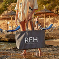 Person holding a gray tote bag with 'REH' branding on a beach.