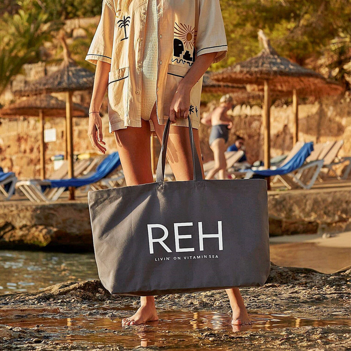 Person holding a gray tote bag with 'REH' branding on a beach.