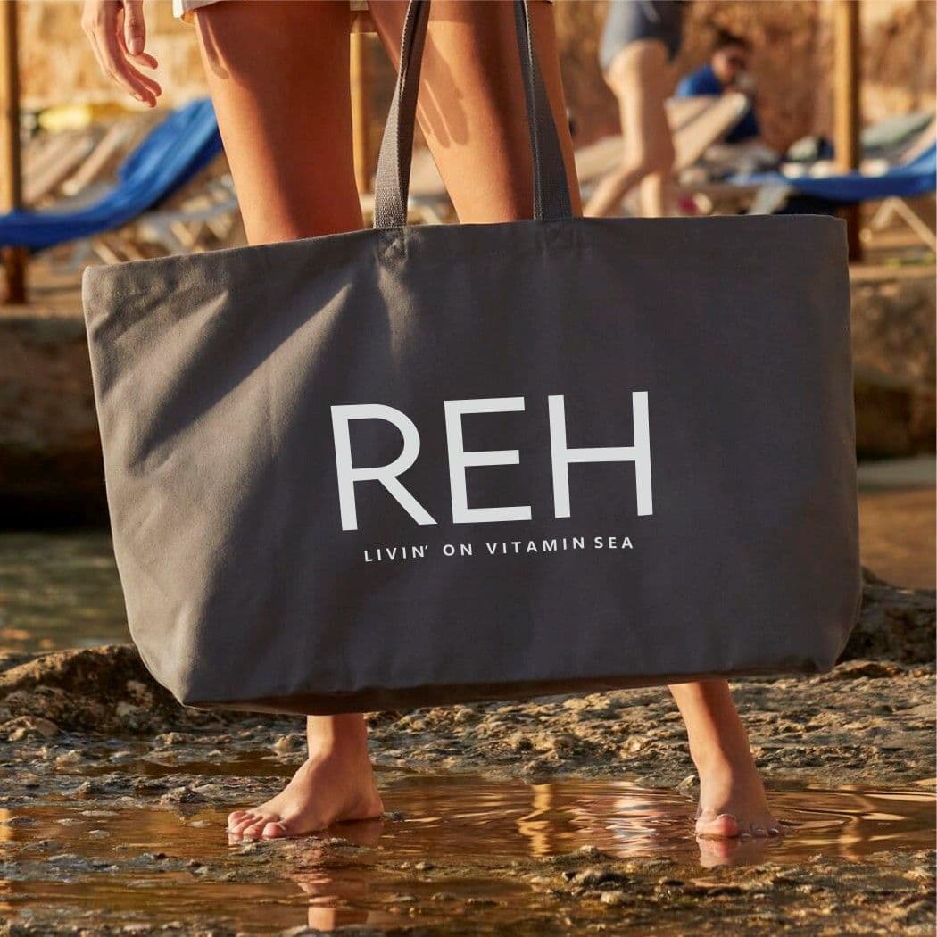 Person holding a large tote bag with 'REH' branding on a beach.