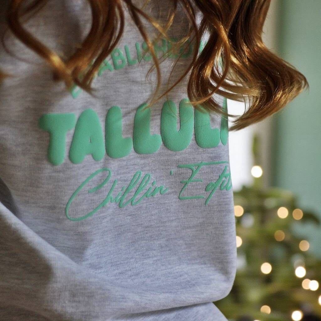 Gray sweatshirt with green text worn by a person, blurred background with lights.