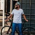 Man wearing a white t-shirt with a bicycle logo, standing next to a black bicycle against a textured wall.