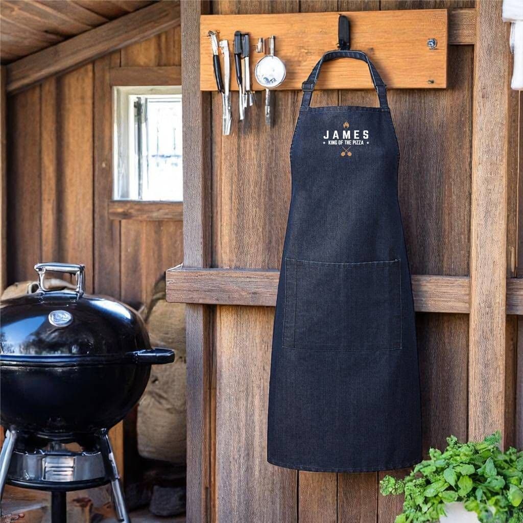 Black apron with 'JAMES' branding hanging on a wooden wall next to a barbecue grill.