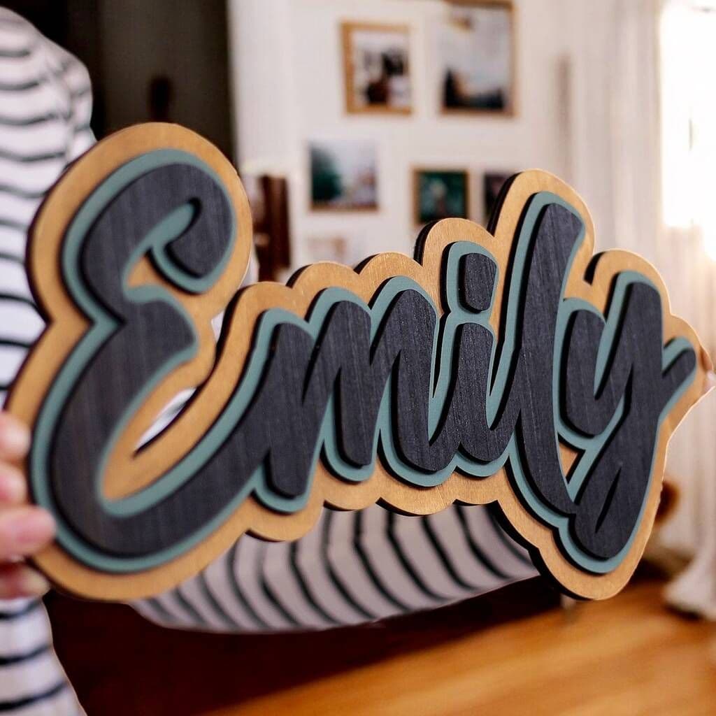 Wooden nameplate with 'Emily' in black and green held by a person in a room with blurred background
