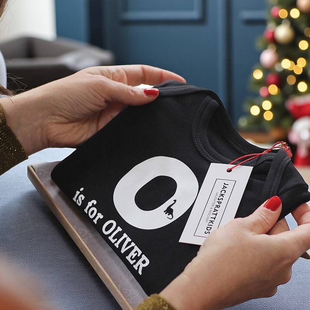 Person holding a black baby romper with 'O is for Oliver' text and a Christmas tree in the background.