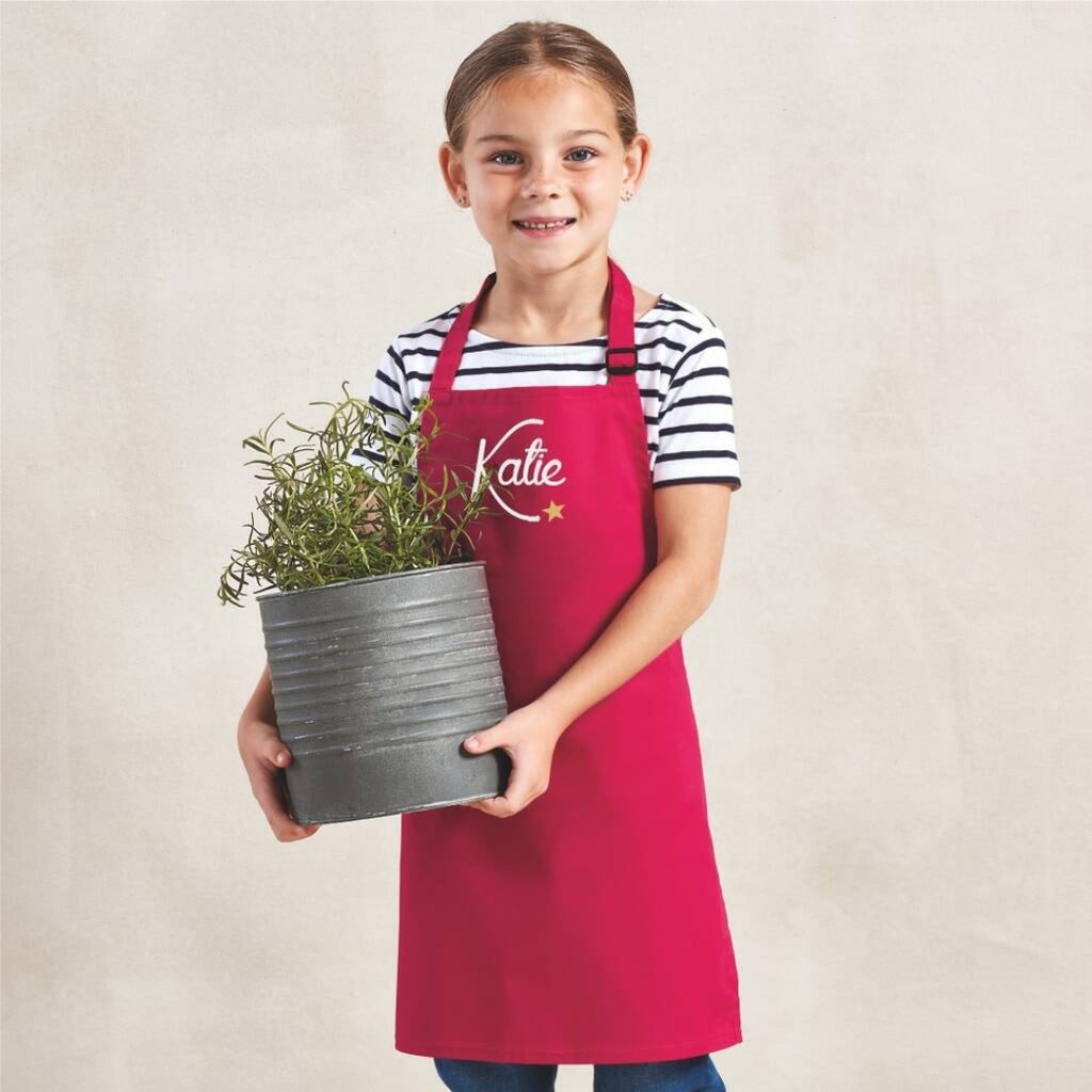 Child wearing a red apron with 'Katie' printed on it, holding a potted plant.