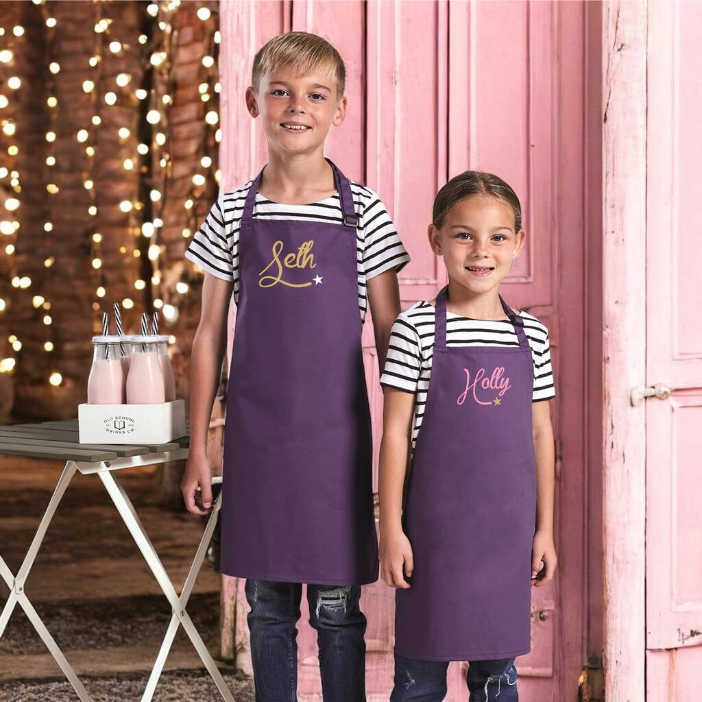 Two children wearing purple aprons with embroidered names, standing in front of a pink door.