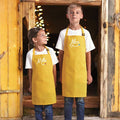 Two children wearing yellow aprons with embroidered names, standing in a rustic wooden doorway.
