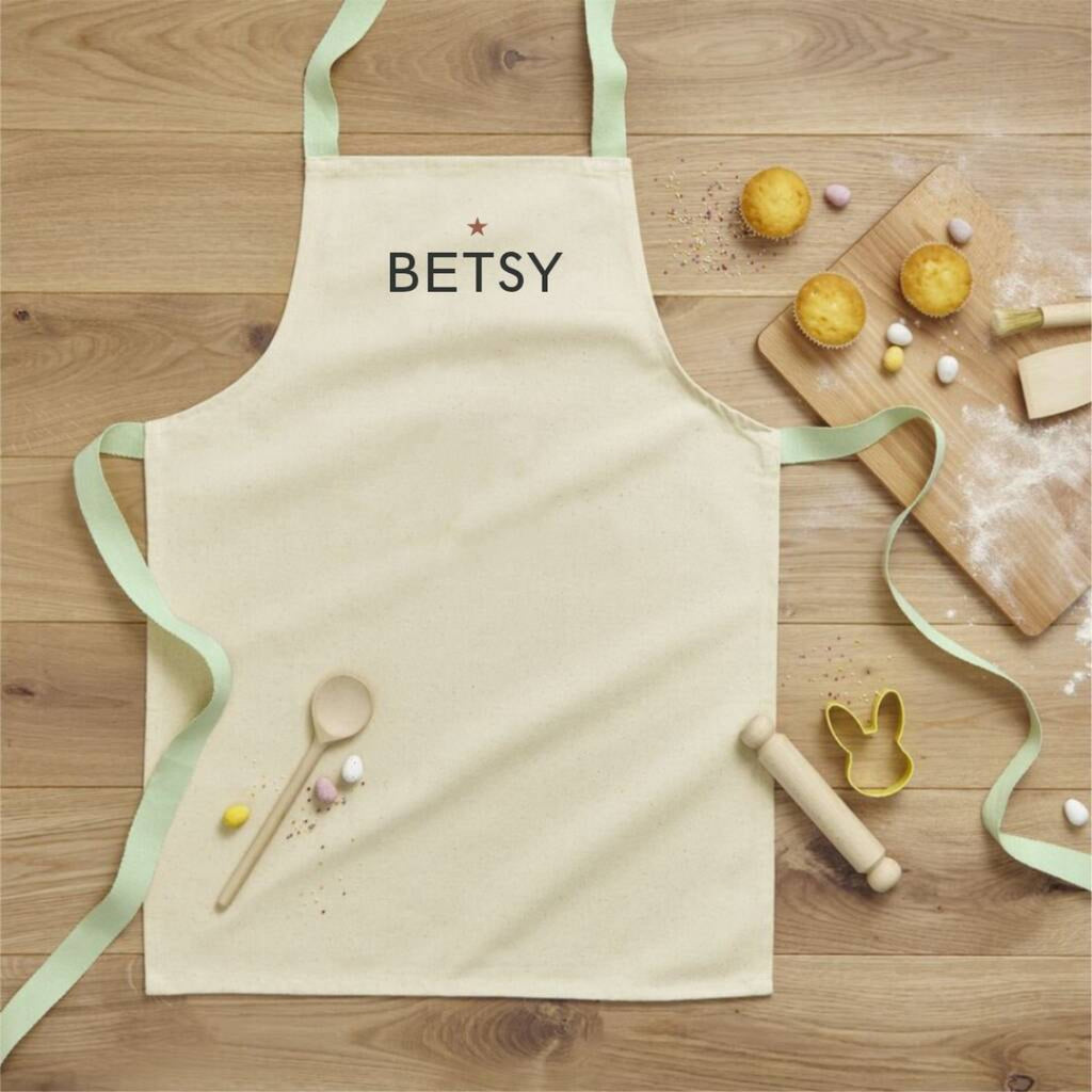 Beige apron with 'BETSY' printed on it, placed on a wooden surface with baking items.