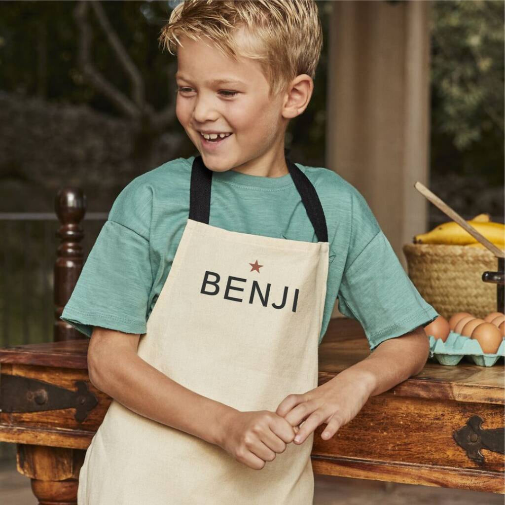 Child wearing a beige apron with 'BENJI' printed on it, standing in a rustic kitchen setting.