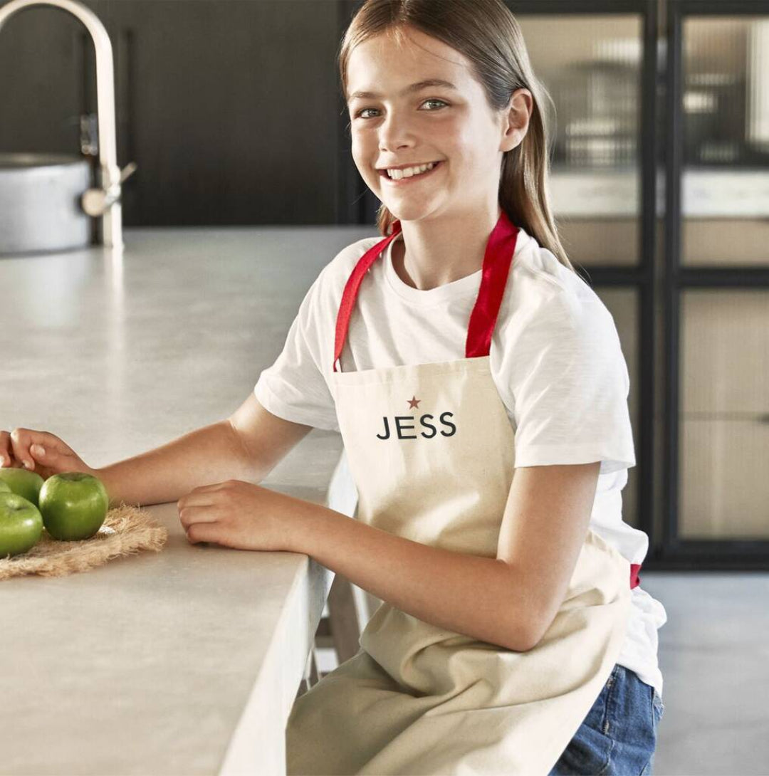 Young girl in a kitchen wearing a beige apron with 'JESS' on it, standing next to green apples.