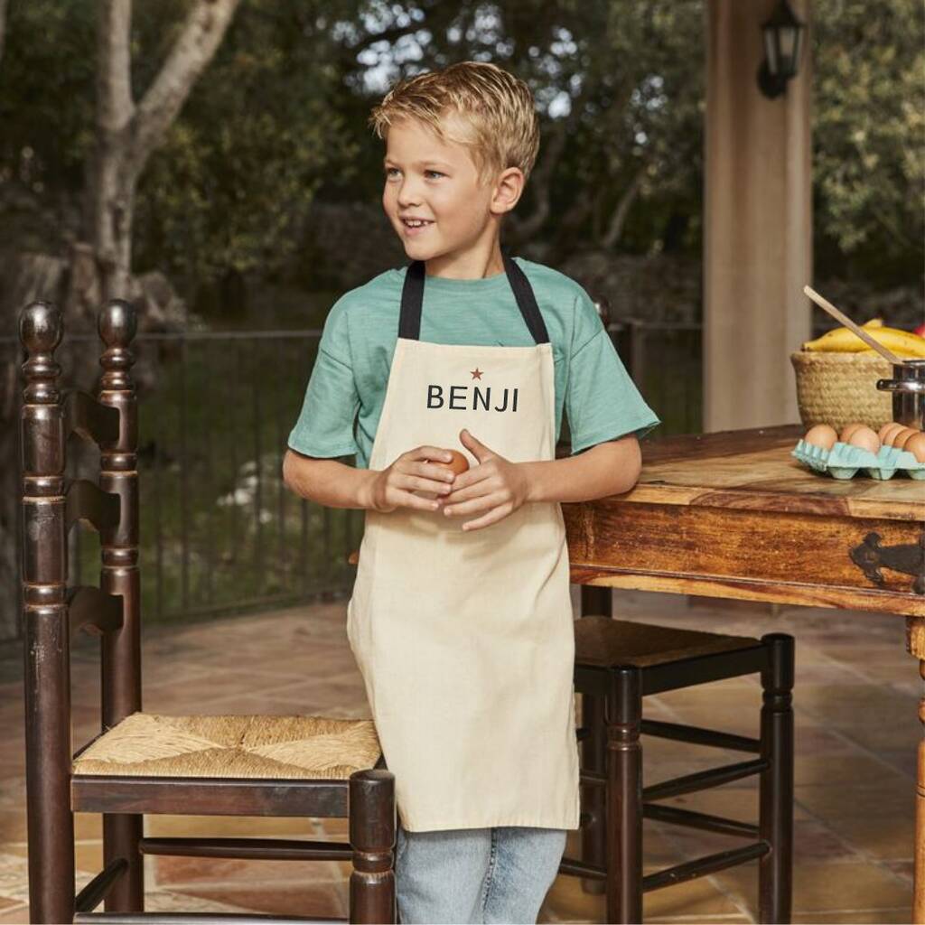 Child wearing a beige apron with 'BENJI' printed on it, standing in a rustic outdoor setting.
