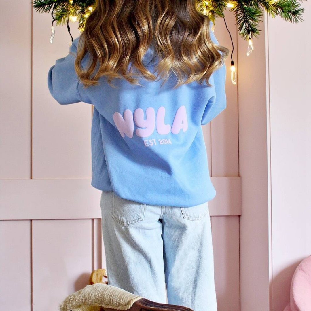 Person wearing a blue puff hoodie with 'Nyla' printed on it, standing in front of a pink wall.
