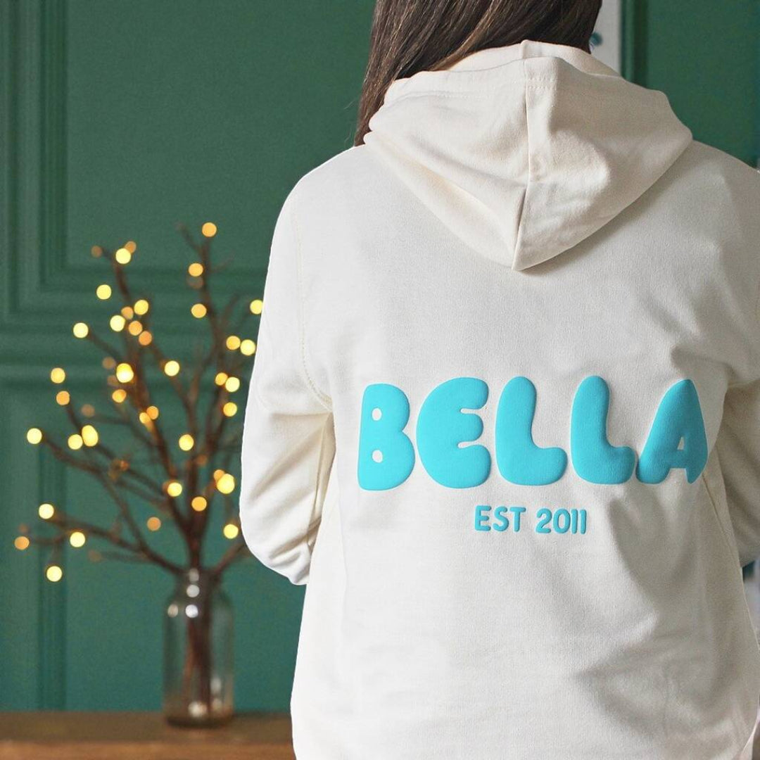 Person wearing a vanilla puff hoodie with 'Bella' and 'Est 2011' text on the back, standing in front of a green wall with decorative lights.