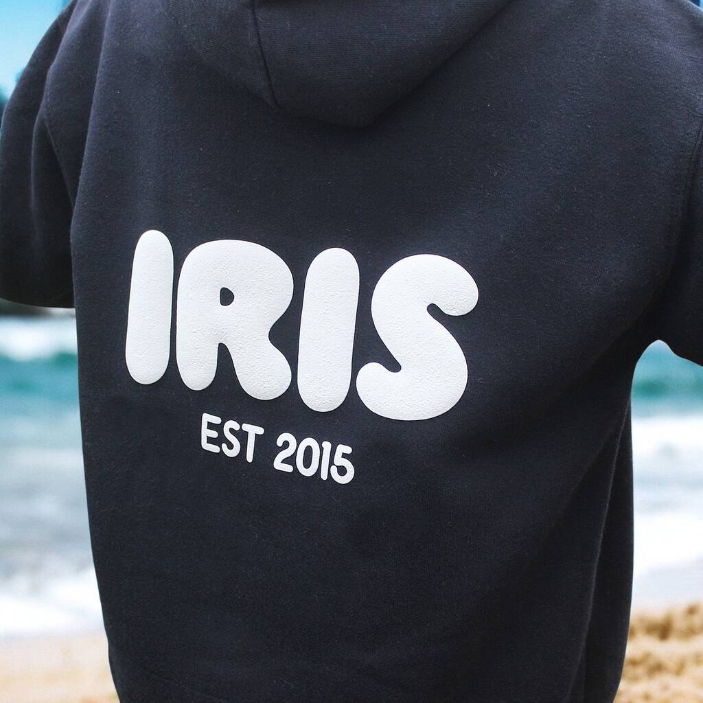 Navyhoodie with 'IRIS EST 2016' text on the back, worn by a person at a beach.