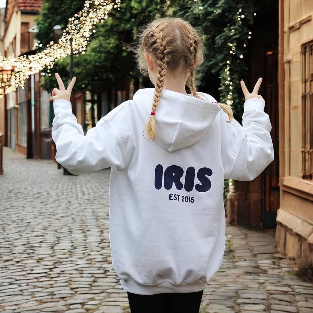 Person wearing a white puff hoodie with 'IRIS EST 2016' on the back, standing on a cobblestone street.