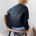 A child seen from behind wearing a black sweatshirt with a gray graphic that reads 'Noah COOL KIDS CLUB OFFICIAL MEMBER ESTABLISHED 2019', sitting on a brown leather stool in a bright indoor setting.