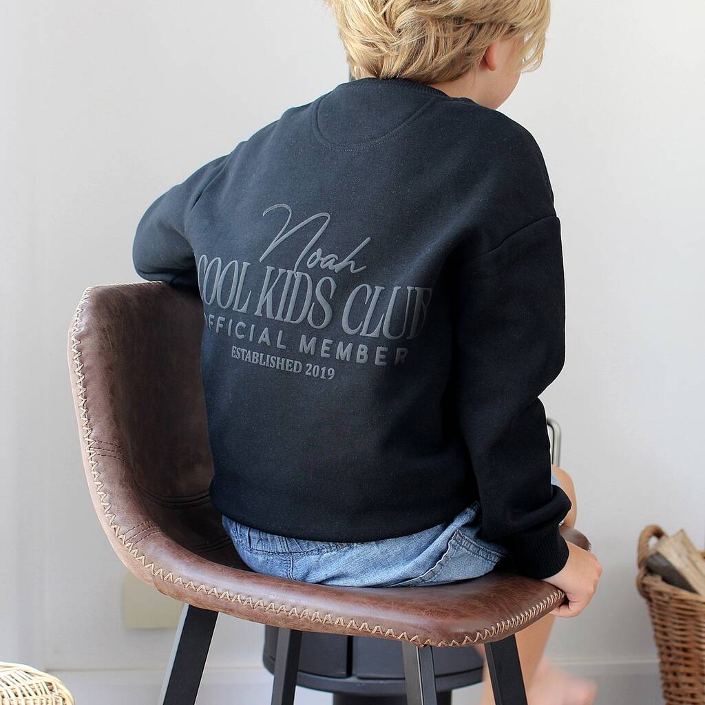 A child seen from behind wearing a black sweatshirt with a gray graphic that reads 'Noah COOL KIDS CLUB OFFICIAL MEMBER ESTABLISHED 2019', sitting on a brown leather stool in a bright indoor setting.