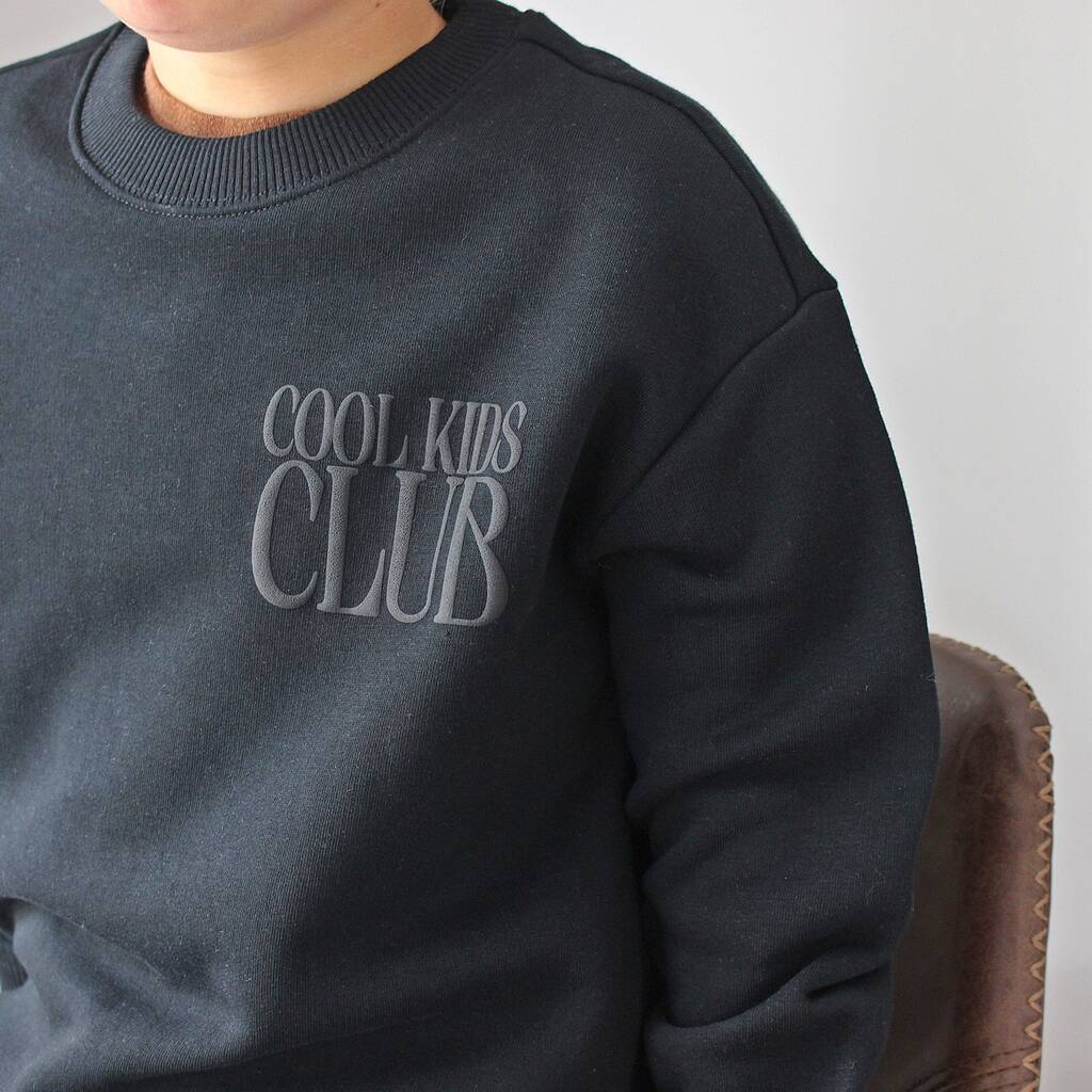A close-up of a person wearing a dark charcoal gray crewneck sweatshirt with 'COOL KIDS CLUB' printed in a matching gray tone on the chest, seated next to a brown leather chair.