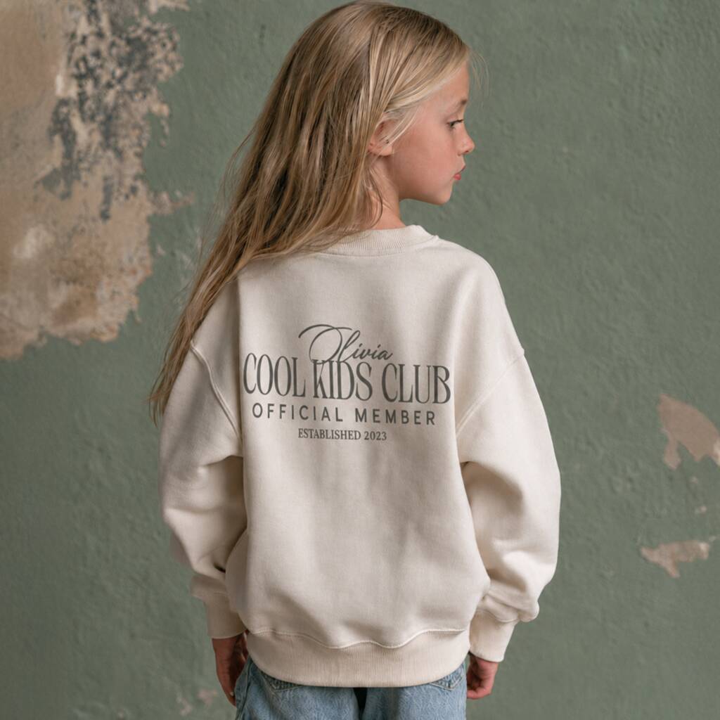 A young girl with long blonde hair is shown from the back, wearing an oversized cream sweatshirt with 'Olivia COOL KIDS CLUB OFFICIAL MEMBER' printed in black on the back. She is standing against a textured sage green wall with peeling paint.