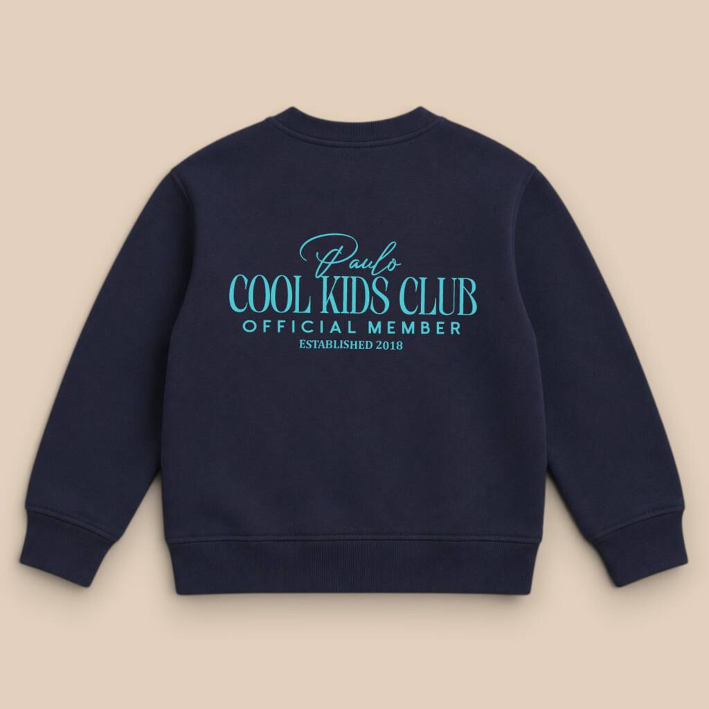 A navy blue crewneck sweatshirt shown from the back, featuring a light blue graphic print that reads 'Paul's COOL KIDS CLUB OFFICIAL MEMBER ESTABLISHED 2019' on a solid beige background.