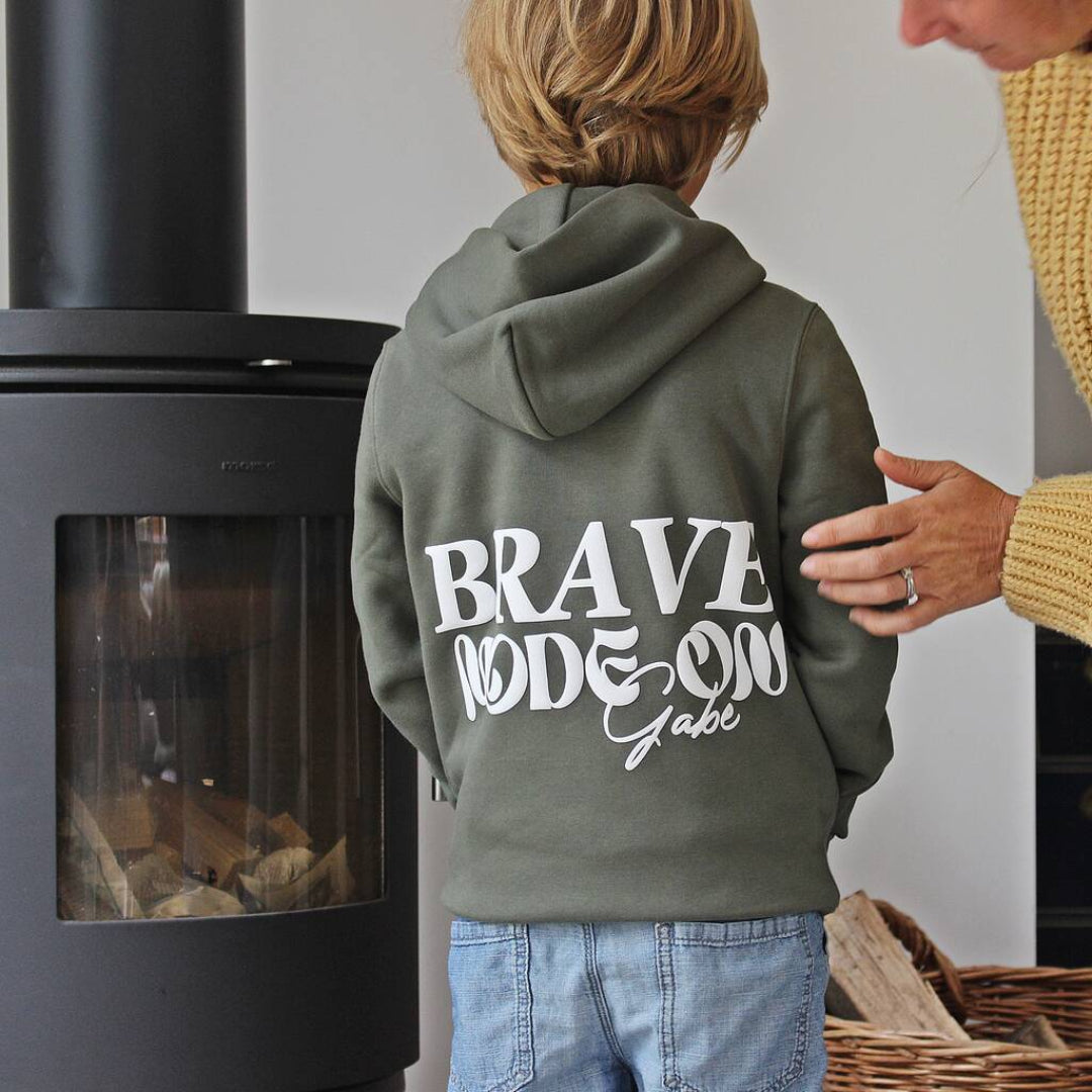 A child viewed from behind wearing an olive green hoodie with 'BRAVE MODE ON Gabe' printed in white across the back, standing indoors next to a black wood-burning stove while an adult hand rests on their shoulder.