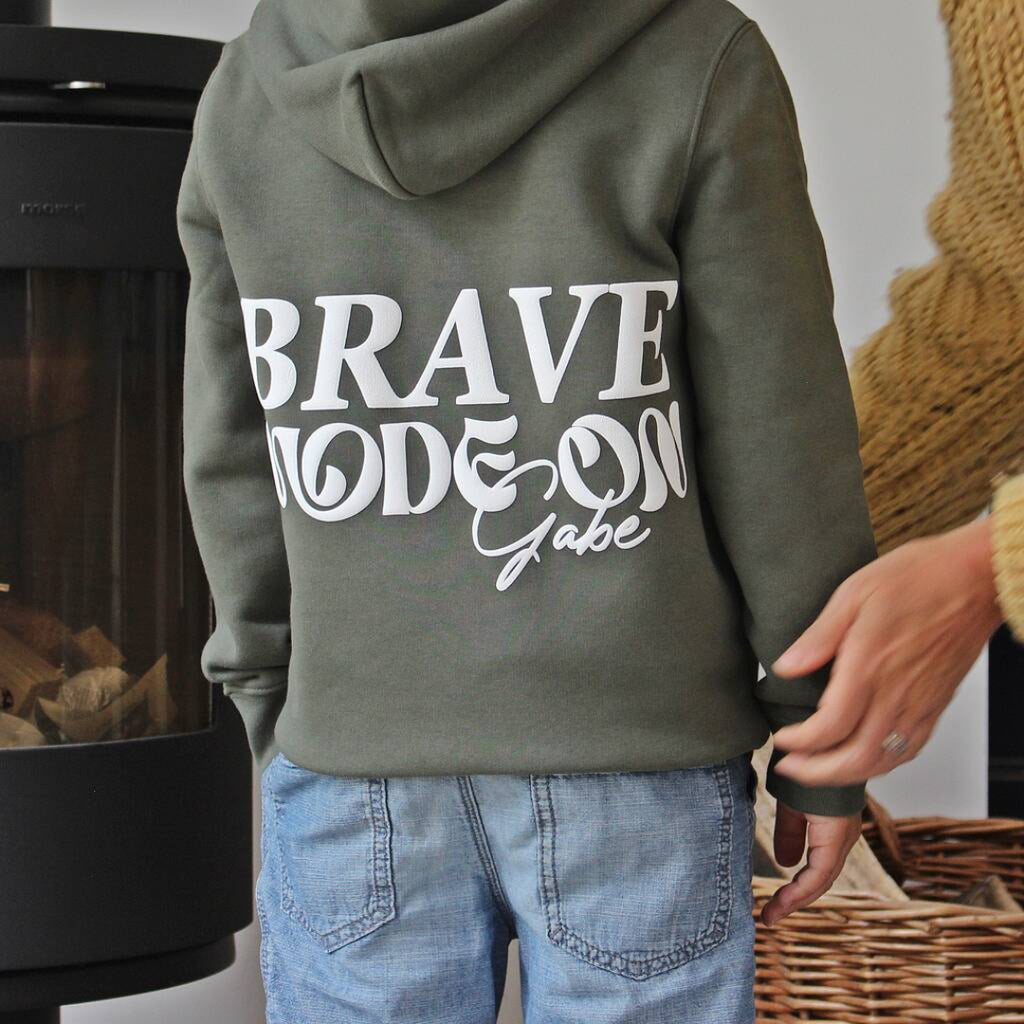 A child wearing an olive green hoodie with 'BRAVE MODE ON Gabe' printed in white on the back, paired with light blue jeans, standing indoors near a fireplace.