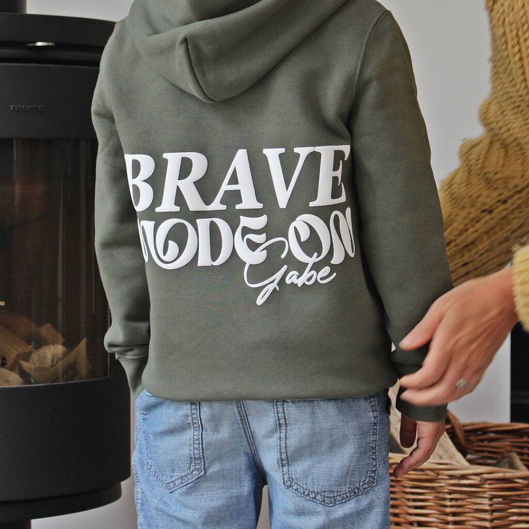 A child wearing an olive green hoodie with 'BRAVE MODE ON Gabe' printed in white on the back, paired with light blue jeans, standing indoors near a fireplace.