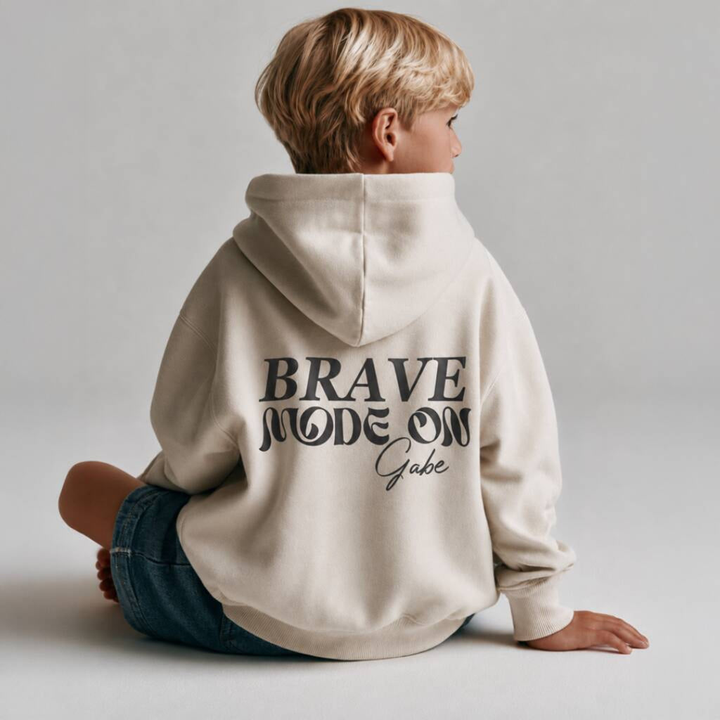 A child wearing a cream-colored hoodie with 'BRAVE MODE ON Gabe' printed in black on the back, sitting against a light gray background.