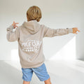 A child wearing a light beige hoodie with 'Pura Smile Club Official Member' and a smiley face graphic printed in white on the back, paired with blue denim shorts, posing with arms outstretched in a bright studio setting.