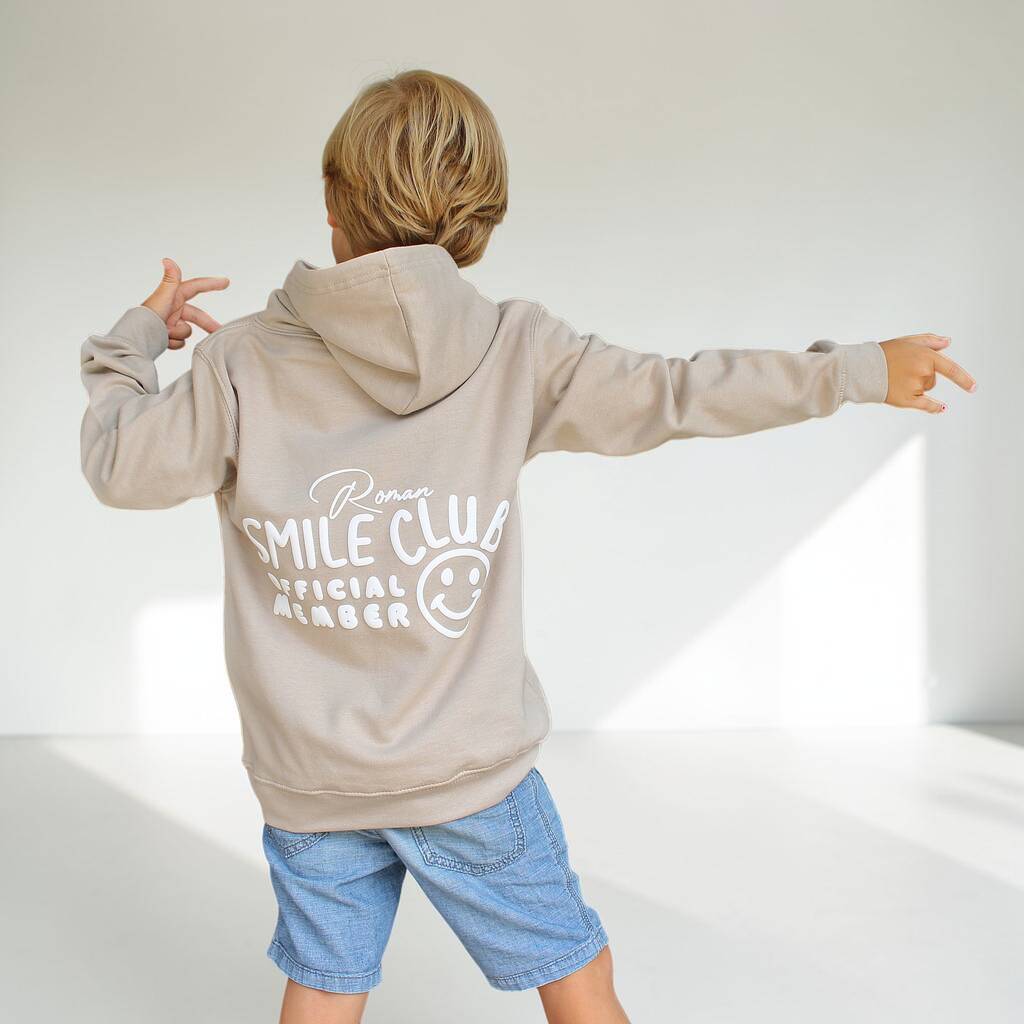 A child wearing a light beige hoodie with 'Pura Smile Club Official Member' and a smiley face graphic printed in white on the back, paired with blue denim shorts, posing with arms outstretched in a bright studio setting.