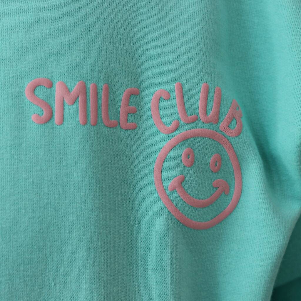 A close-up of a mint green fabric surface with a pink raised 'SMILE CLUB' text logo and smiley face graphic.