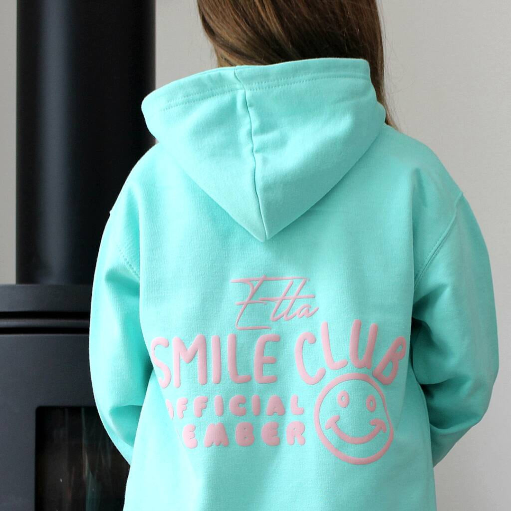 A child wearing a mint green hoodie from the back, featuring a pink embroidered graphic that reads 'Elva SMILE CLUB OFFICIAL MEMBER' with a smiley face, standing indoors next to a dark fireplace.
