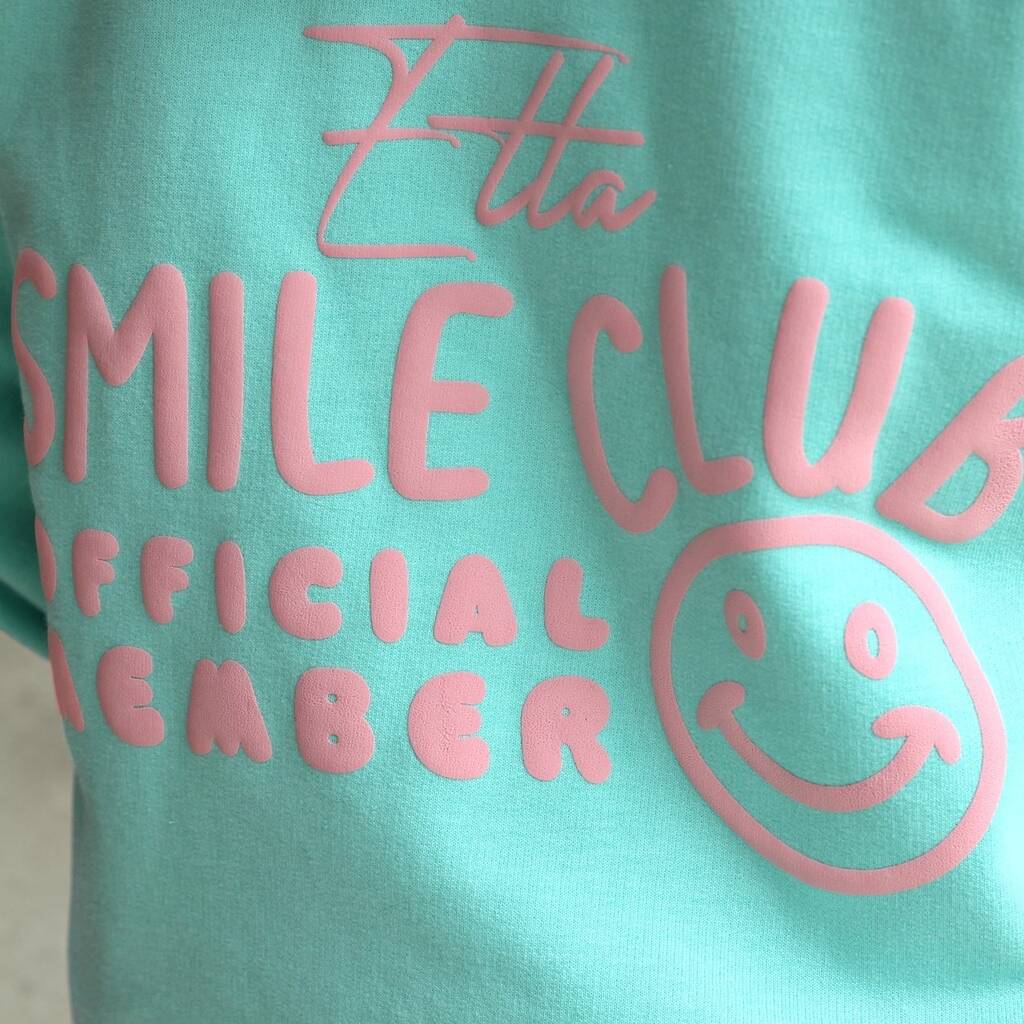 A close-up of a mint green t-shirt featuring a pink raised graphic that reads 'Etta SMILE CLUB OFFICIAL MEMBER' with a smiley face, showing the fabric's 3D texture.