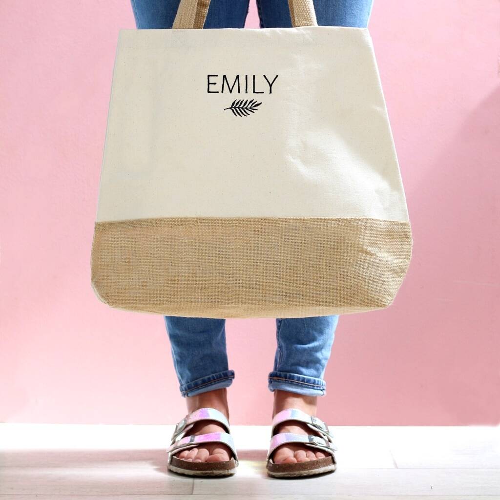 Person holding a beige tote bag with 'EMILY' and a leaf design on a pink background