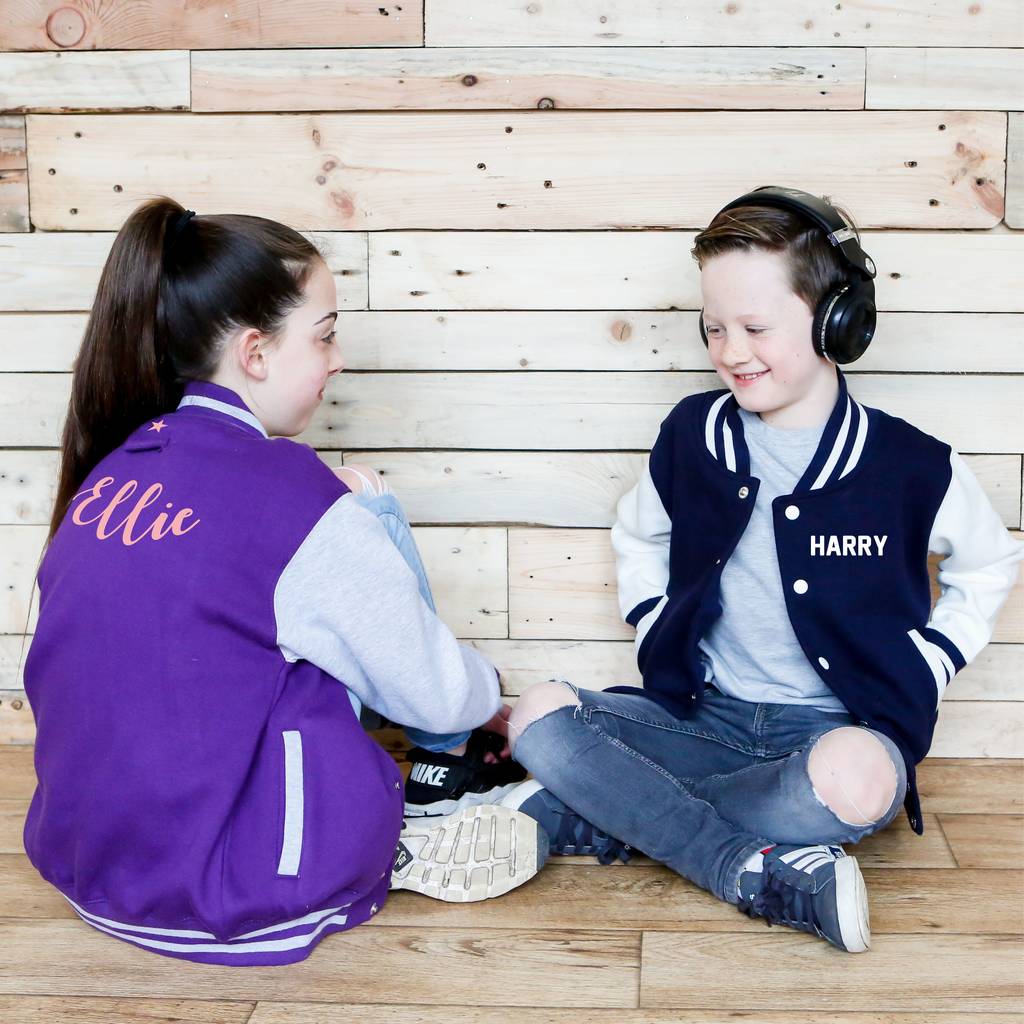 Two children sitting on the floor against a wooden wall, wearing personalized letterman jackets.