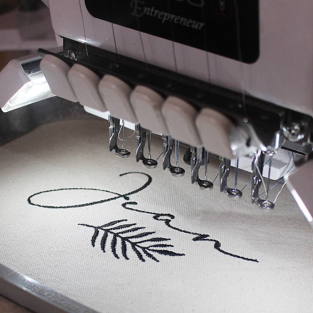 Close-up of an embroidery machine stitching a black script name and leaf design onto white fabric.