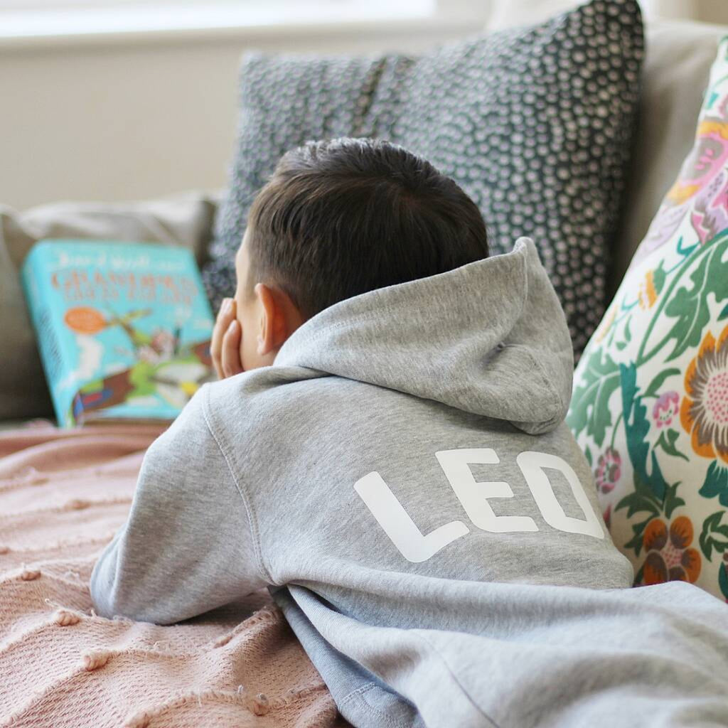 Person wearing a grey onesie with 'LEO' on it, sitting on a couch with colorful pillows and a book.