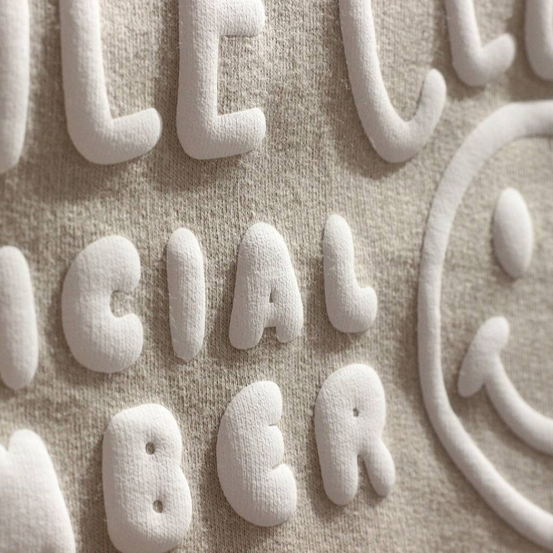 Close-up of a beige textured fabric with raised white letters spelling part of a phrase, alongside a white smiley face outline.