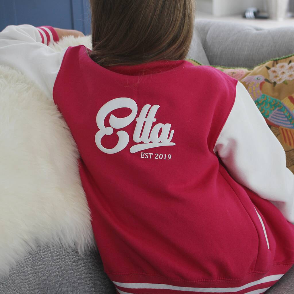 A person sitting on a gray sofa wearing a red and white varsity-style jacket, viewed from behind. The back of the jacket has a large white script logo reading 'Etta' with 'EST 2019' underneath.