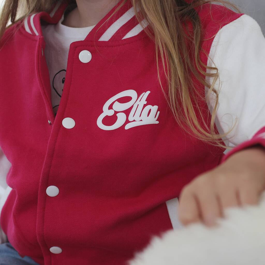A close-up of a child wearing a red varsity-style jacket with white sleeves, white snap buttons, and a white script logo reading 'Elta' on the chest.