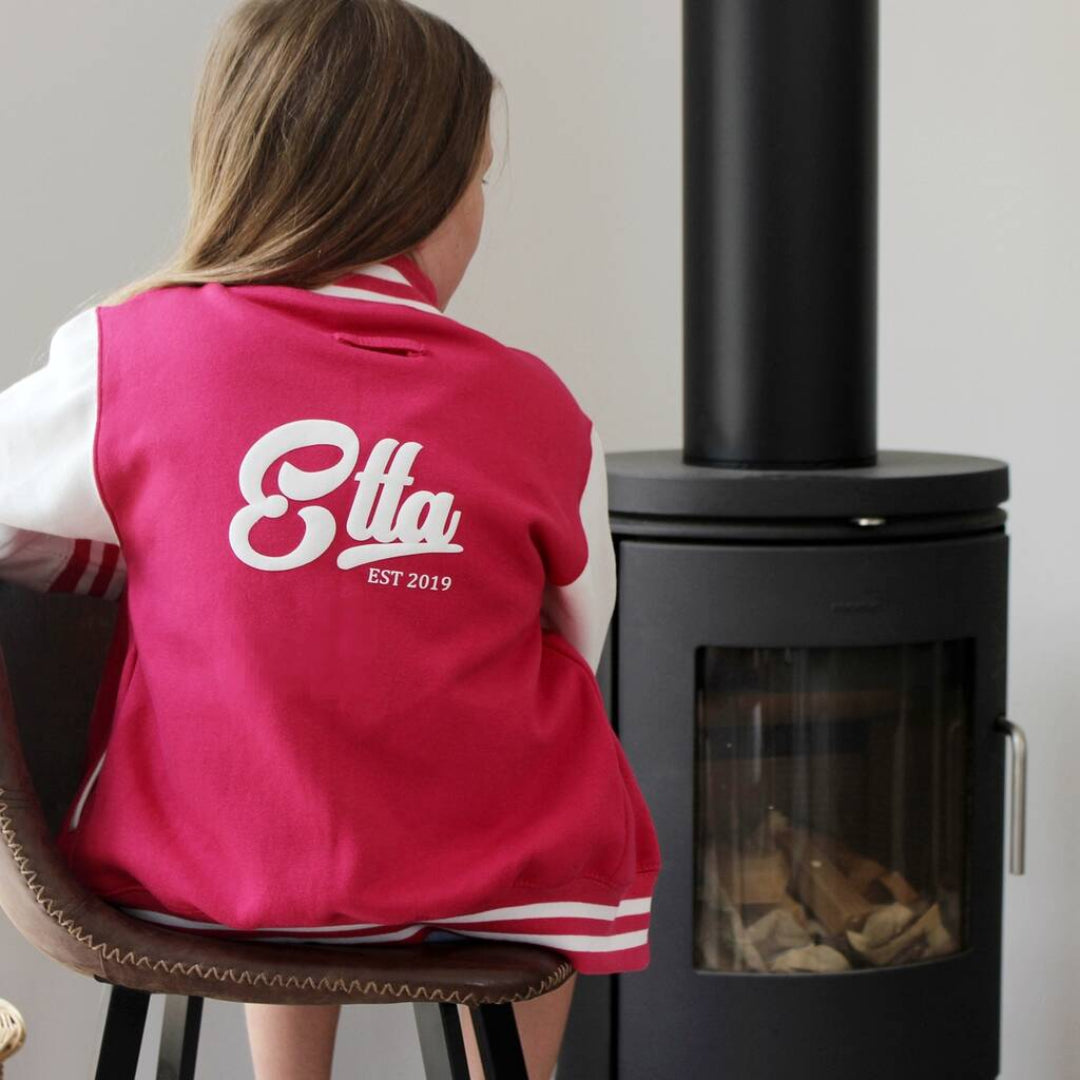 A person sitting on a chair, viewed from behind, wearing a bright pink varsity jacket with white sleeves and white lettering that reads 'Etta EST 2019' on the back. The person is seated in front of a black wood-burning stove.