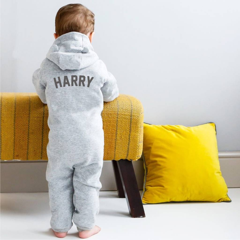 Child wearing a grey onesie with 'HARRY' printed on it, standing in a room with yellow cushions and a white wall.