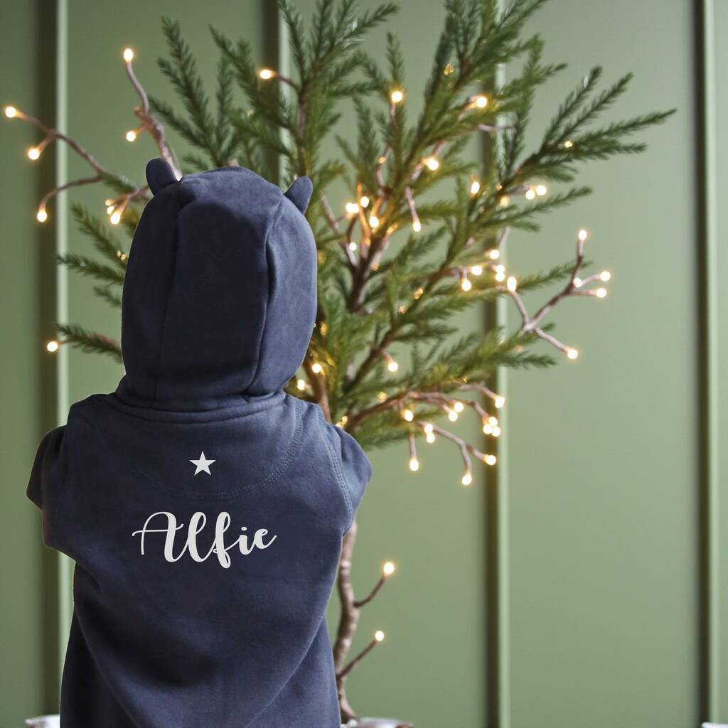 Child wearing a navy blue onesie with 'Alfie' printed on the back, standing in front of a decorated Christmas tree.