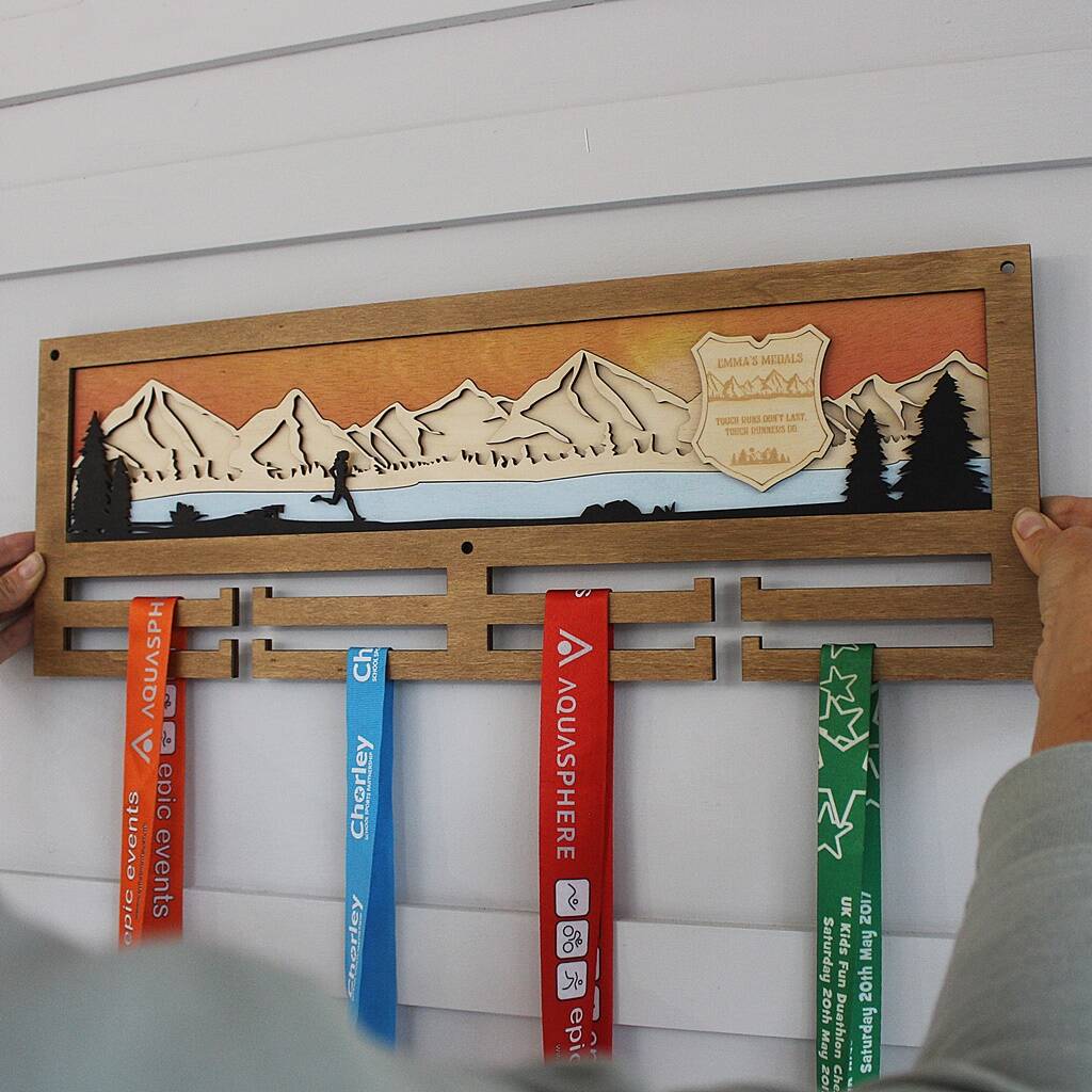 Wooden medal holder with engraved mountain scene and ribbons on a white wall