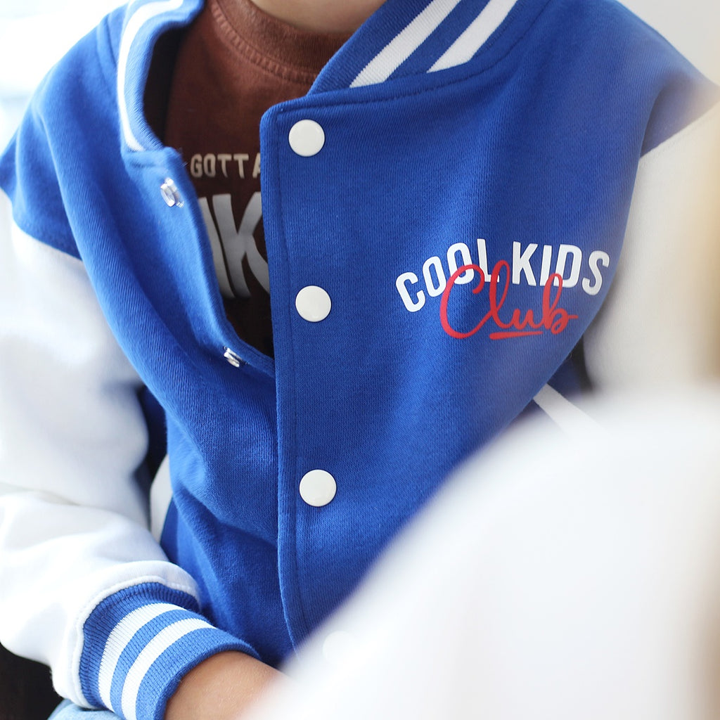 A close-up of a child wearing a blue and white varsity-style jacket with 'COOL KIDS Club' embroidered on the chest, layered over a brown shirt.