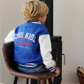 A child seen from behind wearing a blue and white varsity jacket with 'COOL KIDS Club SINCE 2021' printed on the back, sitting on a brown leather chair in front of a black stove.