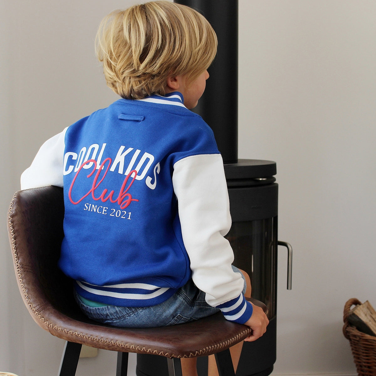 A child seen from behind wearing a blue and white varsity jacket with 'COOL KIDS Club SINCE 2021' printed on the back, sitting on a brown leather chair in front of a black stove.