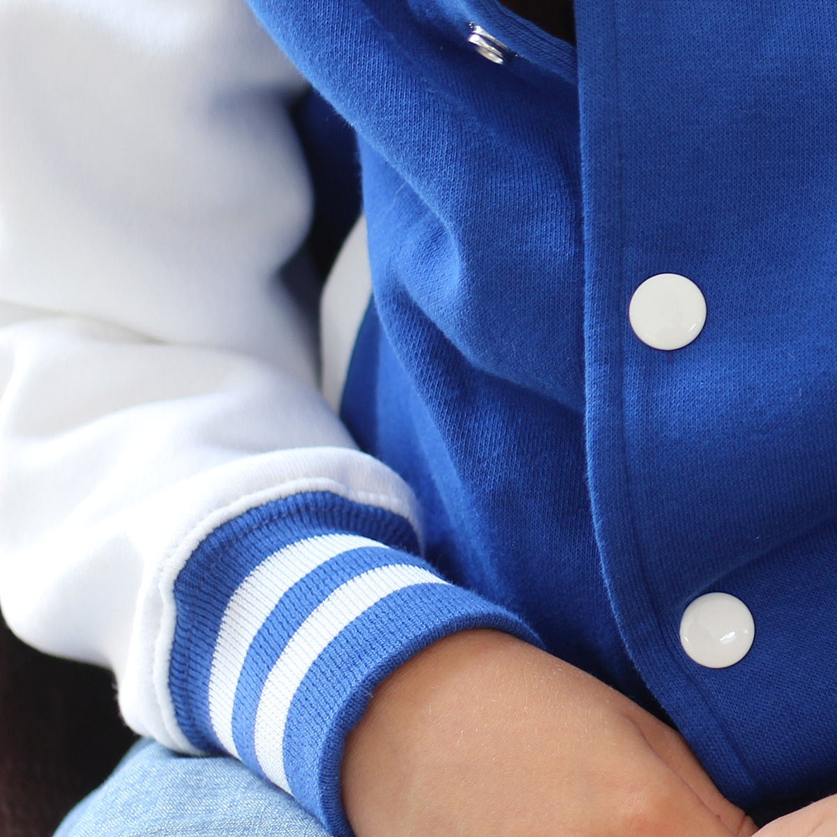Close-up of a blue and white varsity-style jacket worn by a child, showing the ribbed blue-and-white sleeve cuff and white snap buttons.